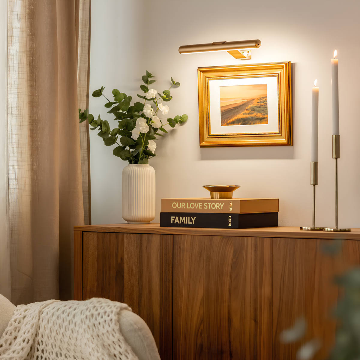 Lampe de tableau dorée au-dessus d'un cadre, à côté de bougeoirs sur une commode