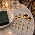 Ménagère couverts doré mat à côté d'une assiette blanche sur une table en marbre